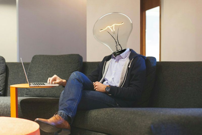 Homme assis sur un canapé, qui est sur son portable et qui a une ampoule à la place de la tête